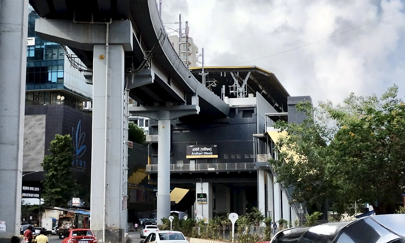 Andheri West metro station viewed from New Link Road, with its station sign, elevated tracks and street traffic.